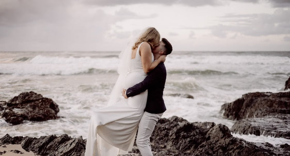 A newlywed couple shares a passionate kiss while being lifted on a rocky beach, embodying the perfect start to a romantic stay in Byron Bay.
